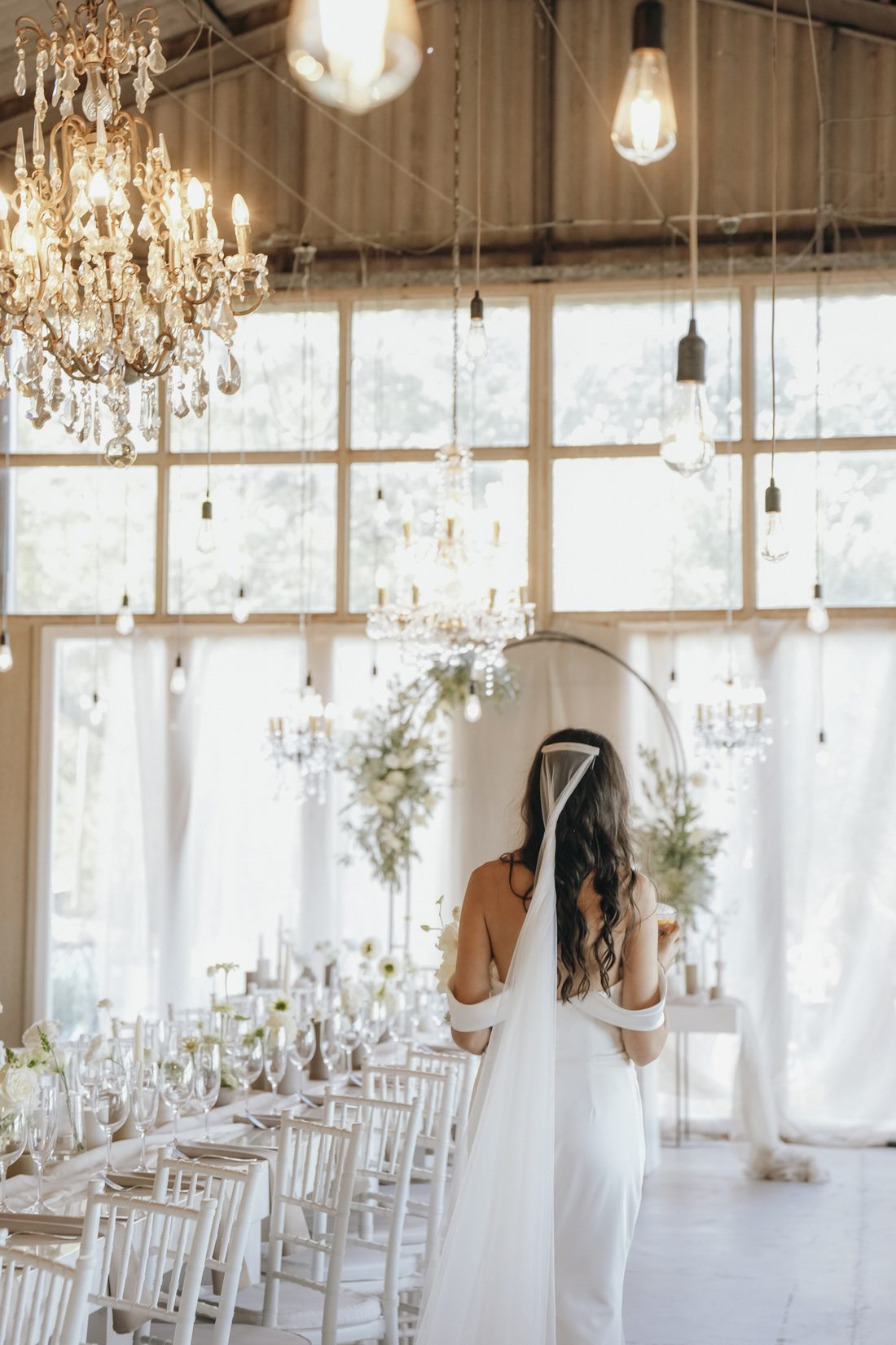 A bride in the barn beneath the chandeliers, long tables set for the celebration