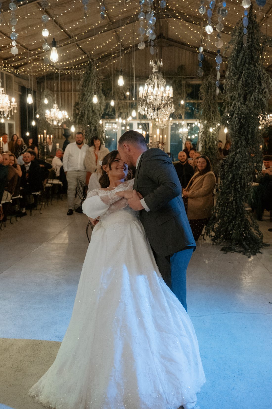 A couple dancing beneath the chandeliers and fairy lights in the barn