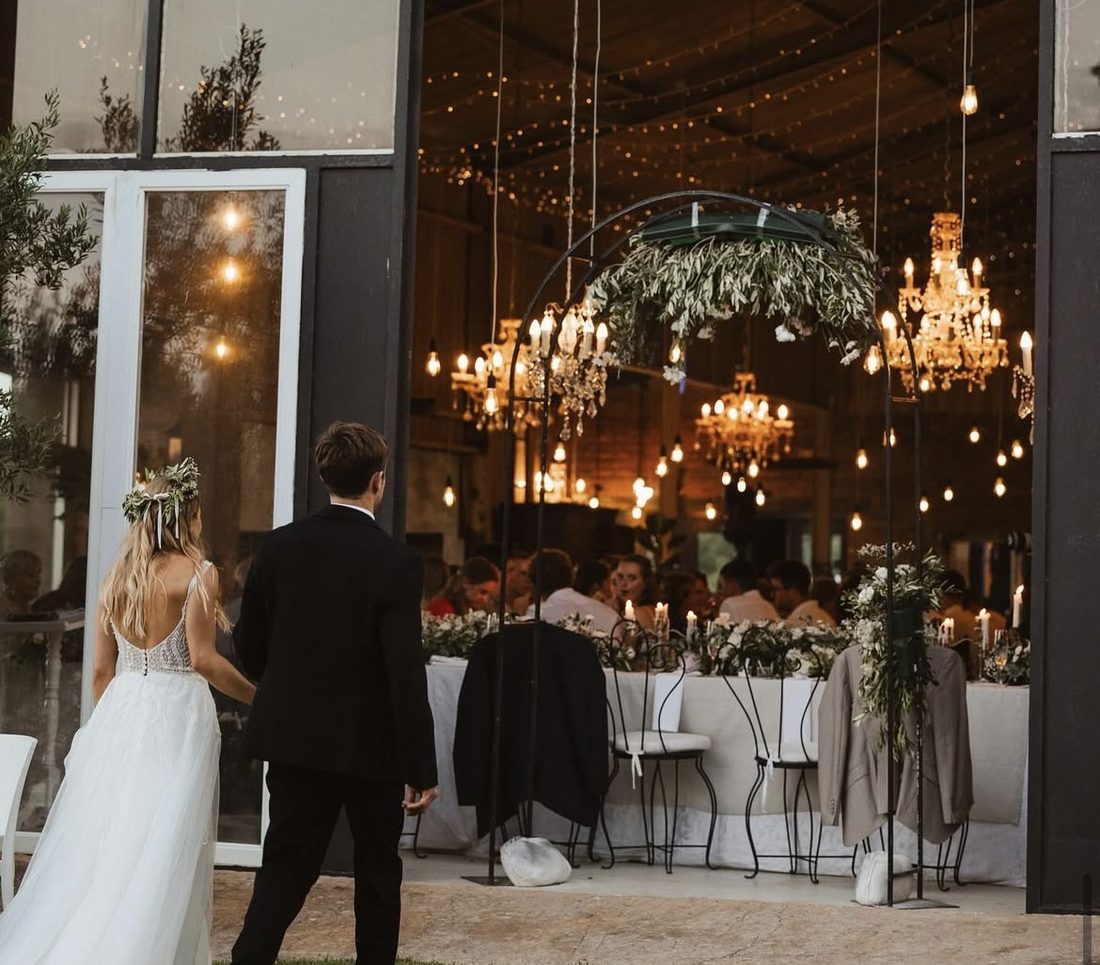 A couple stepping into the barn as their guests are seated beneath the chandeliers