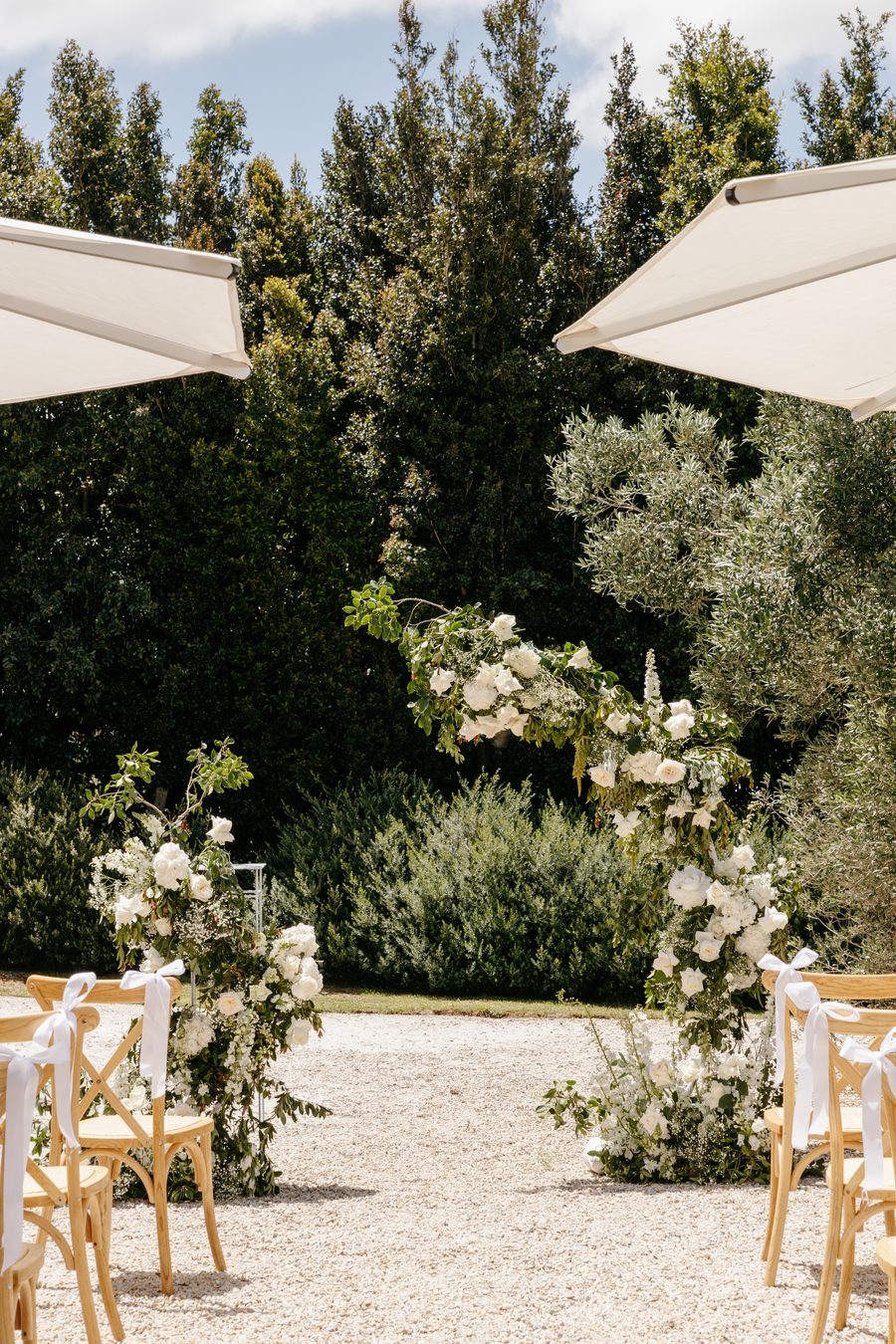 A garden ceremony aisle framed by white florals, cross back chairs and white umbrellas